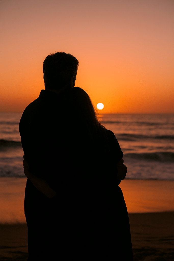 Silhouette of a man with short gray hair holding his wife from behind as they stand on a beach watching a vivid orange sunset over the ocean.